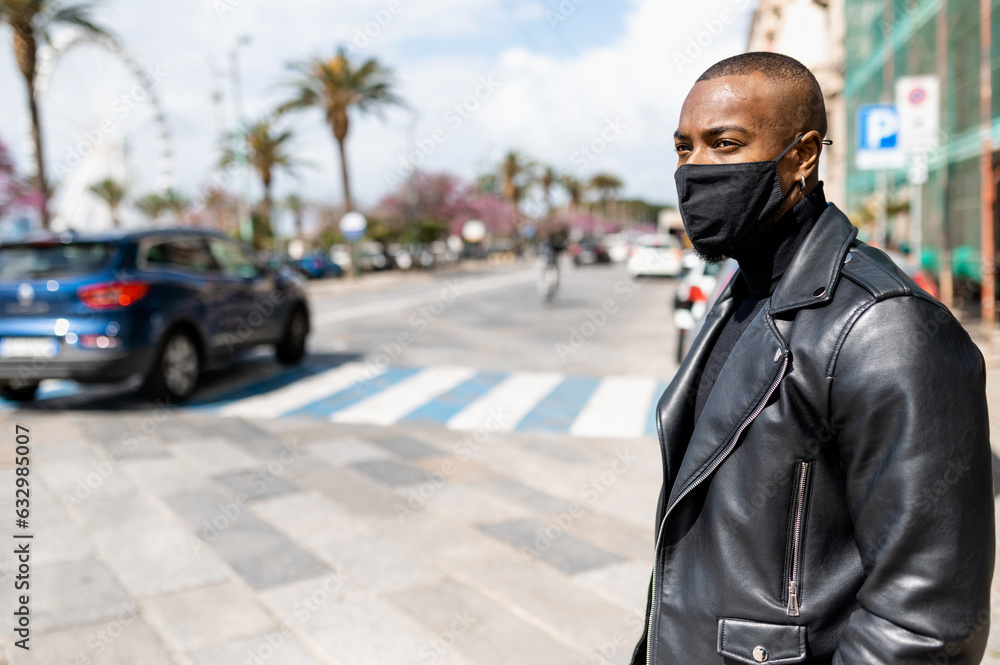 Side view thoughtful African American male in face mask and leather jacket standing on roadside in sunny tropical city and looking away
