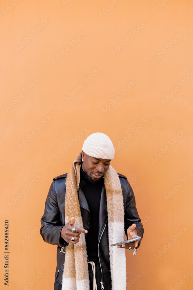 Fototapeta premium Happy stylish African American male in leather jacket hat and scarf browsing mobile phone while standing against brown wall