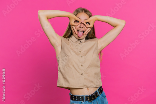 Funny comical playful young woman making silly facial expressions and grimacing, fooling around, showing tongue, idiotic expression. Girl isolated alone on pink studio background. People lifestyles