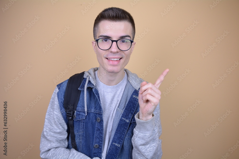 Smiling Student Pointing Upward. Hmiling Young Man in Glasses, Wearing Backpack on Isolated Background Smiling Student Pointing Upward. Hmiling Young Man in Glasses, Wearing Backpack on Isolated Background