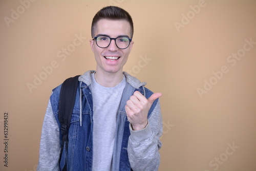 Smiling Student Giving Thumbs Up. Smiling Young Man in Glasses, Wearing Contrast Sleeves and Backpack on Solid Background. Thumb Up Gesture.