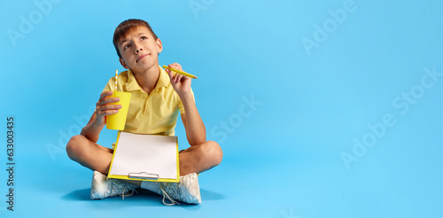 Little school boy, child sitting on floor with notebook, doing homework against blue studio background. Concept of childhood, kids emotions, education, remote school. Banner. Copy space for ad