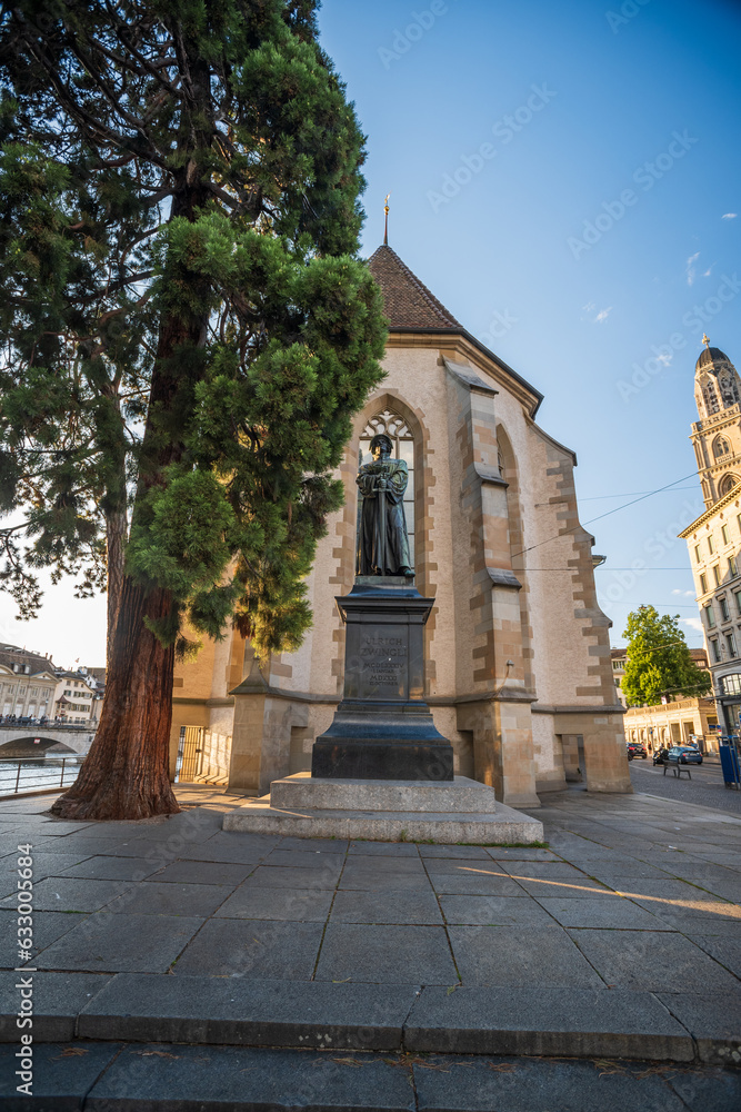 03-08-2023  Zurich city Switzerland. Wide-angle view of Wasserkirche Zürich or Water Church. Setting sun in the background, large tree next to bronze statue