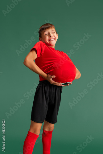 Smiling, cheerful little boy, child in sportswear holding ball under t-shirt against green studio background. Concept of childhood, kids emotions, sportive lifestyle, action, hobby, ad © master1305