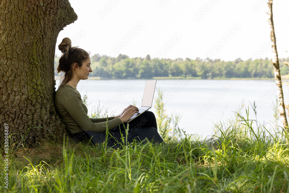 A young and talented female freelancer finds herself in a blissful paradise, working with her laptop while surrounded by the breathtaking view of a serene forest and a tranquil lake. This idyllic