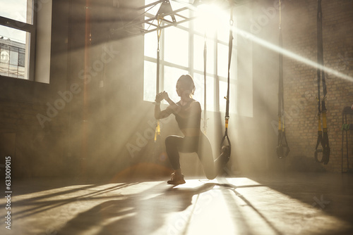 Deep squat. Full-length of young athletic woman in sports clothing training legs with trx fitness straps in the gym photo