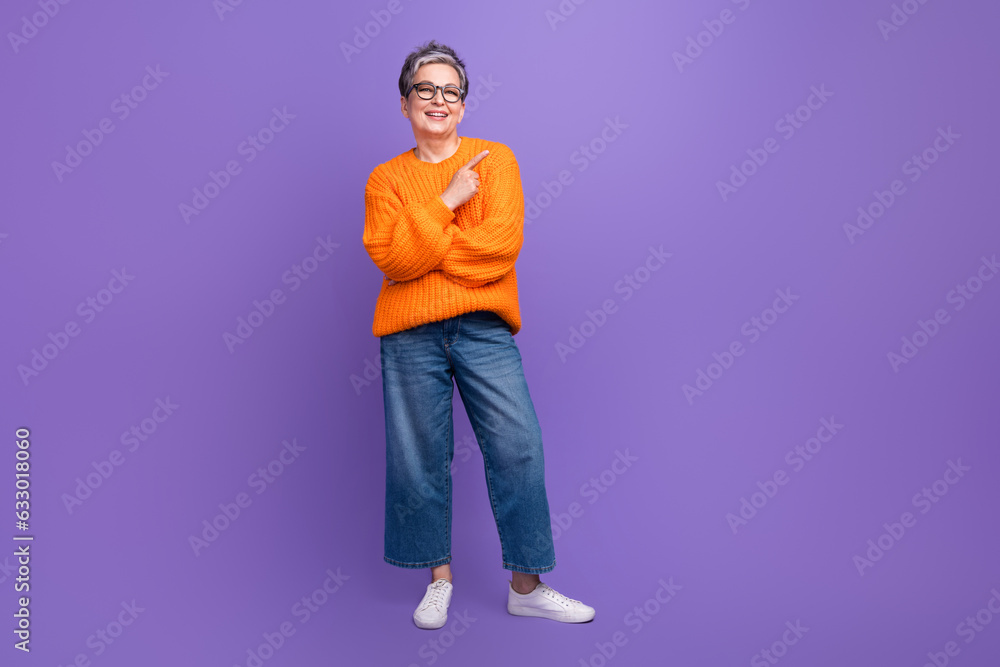 Full length body photo of retired woman wear orange sweater jeans direct finger empty space advert isolated on purple color background Full length body photo of retired woman wear orange sweater jeans direct finger empty space advert isolated on purple color background