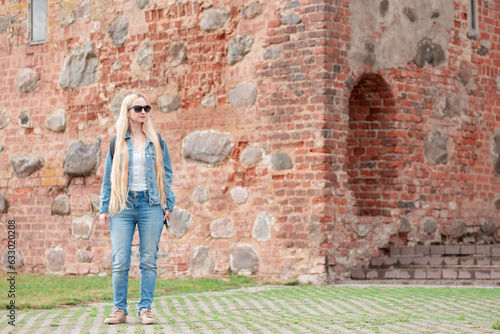 Blonde girl in sunglasses with a backpack in jeans clothes on the background of a brick building. © 7707601