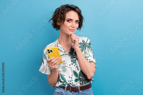 Portrait of minded smart woman with stylish hairdo wear print shirt hand on chin holding smartphone isolated on blue color background