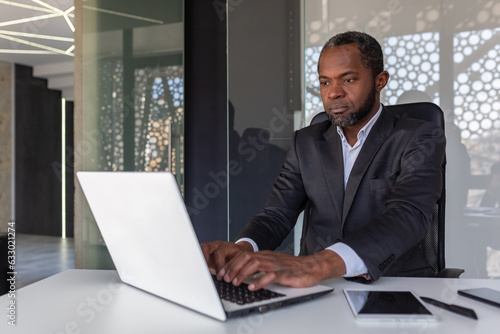 Serious mature businessman working on laptop inside office at workplace, african american boss thoughtful in business suit, successful experienced investor banker.