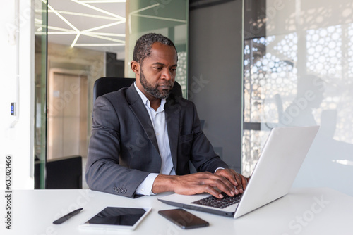 Serious mature businessman working on laptop inside office at workplace, african american boss thoughtful in business suit, successful experienced investor banker.
