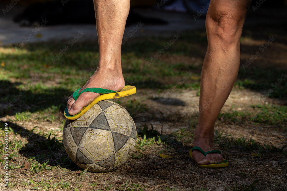 Mulher com um pé em cima de uma bola de futebol, calçada com chinelos verde e amarelo em campo de várzea.