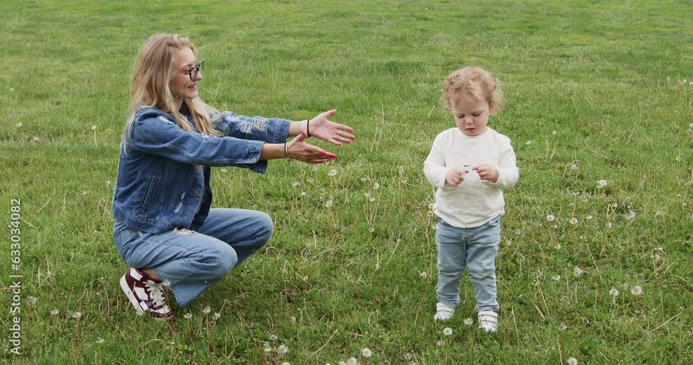 Young mother plays with toddler son on dandelion meadow in city park. Boy holds dandelions and runs to hug mother sitting on grass Young mother plays with toddler son on dandelion meadow in city park. Boy holds dandelions and runs to hug mother sitting on grass