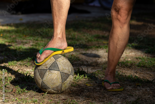 Mulher com um pé em cima de uma bola de futebol, calçada com chinelos verde e amarelo em campo de várzea. photo