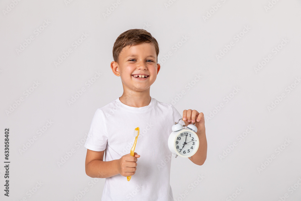 Morning concept. Close-up portrait of a happy little boy with alarm clock and toothbrush on white background. A child in a white T-shirt brushes his teeth on his own. Space for text Morning concept. Close-up portrait of a happy little boy with alarm clock and toothbrush on white background. A child in a white T-shirt brushes his teeth on his own. Space for text