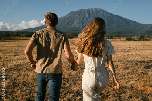 A young couple  a man and a woman  of European appearance  run through the field  holding hands  rear view  and enjoy the view of Mount Agung Volcano  Bali.