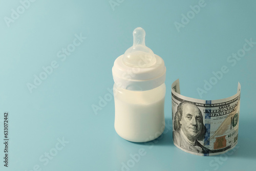 Baby bottle with formula or milk, next to $100. The concept of expensive artificial nutrition for babies. Blue background. Copyspace.