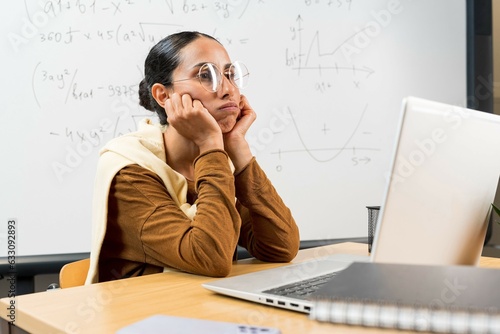 Portrait of exhausted stressed young teacher suffering, sitting at desk near whiteboard, thinking about problems. Arabian girl puts her head on her hands, looks into the distance thoughtfully