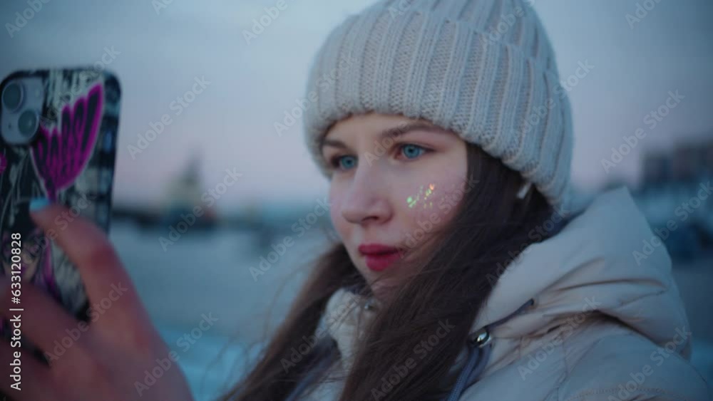 A young caucasian girl with red lipstick, blue eyes and glitter on her face in a winter jacket, long hair and a white hat takes pictures of winter sea and hills on smartphone with multi-colored case A young caucasian girl with red lipstick, blue eyes and glitter on her face in a winter jacket, long hair and a white hat takes pictures of winter sea and hills on smartphone with multi-colored case