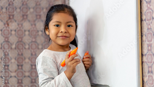 Niña latina sonriente y feliz, escribiendo en la pizarra de clases con plumones de colores. Espacio para texto.