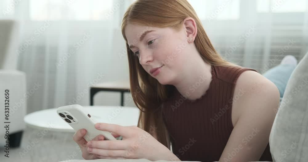 A bored Caucasian teenage girl is seen lying on a sofa, casually browsing her phone. Her facial expression and body language convey a sense of disinterest and ennui. A bored Caucasian teenage girl is seen lying on a sofa, casually browsing her phone. Her facial expression and body language convey a sense of disinterest and ennui.