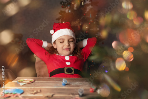 6 yearold girl in Santa Claus costume dreaming with closed eyes at table with decor for Christmas tree