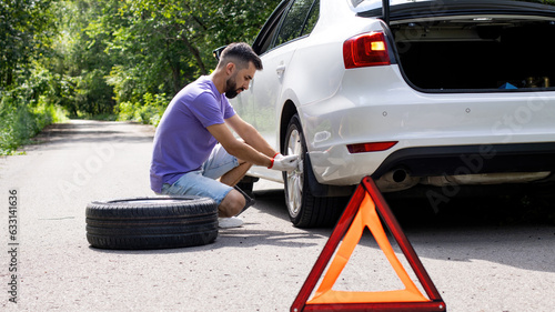 Installing or replacing a car's spare wheel on the side of the road or the edge of the road. Young man in a purple T-shirt and shorts is tightening the nuts and bolts of the wheel. Summer.