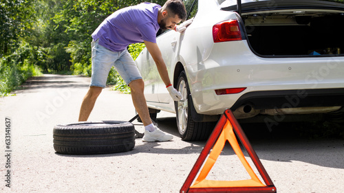 Installing or replacing a car's spare wheel on the side of the road or the edge of the road. Young man in a purple T-shirt and shorts is tightening the nuts and bolts of the wheel. Summer.