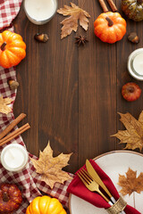 Autumn-themed decor concept. Top view vertical shot of plate, cutlery, napkin, tablecloth, pumpkins, cinnamon sticks, candle, dry leaves on brown wooden table background with space for advert or text