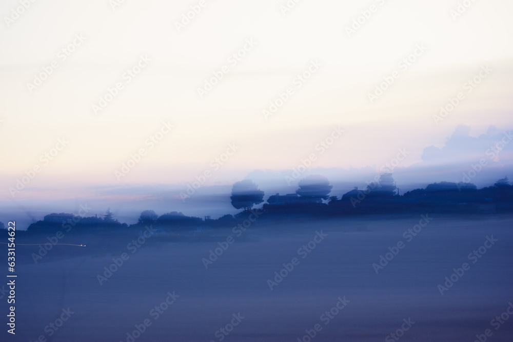 Morning fog in a meadow, field. Blue abstract scenic background with blurred and smeared trees on a horizon against the sky. Beautiful calm landscape with the hills. Autumn sunrise.