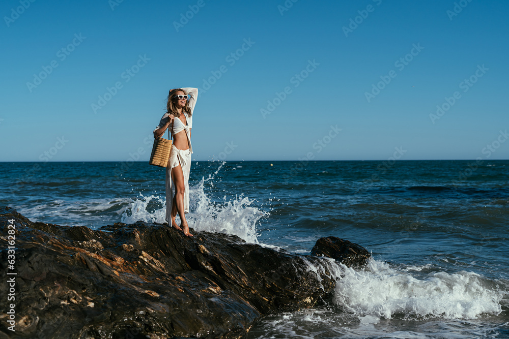 A beautiful girl in white glasses and white clothes with a bag in her hands stands on the stones on the seashore