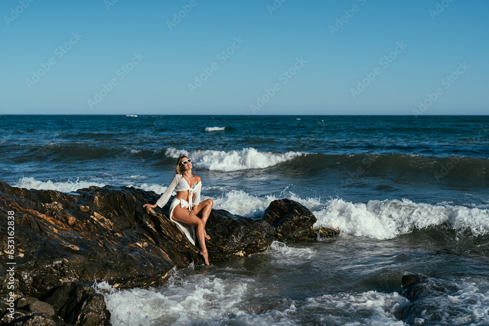 A beautiful girl in white glasses and white clothes sits on the rocks on the seashore A beautiful girl in white glasses and white clothes sits on the rocks on the seashore