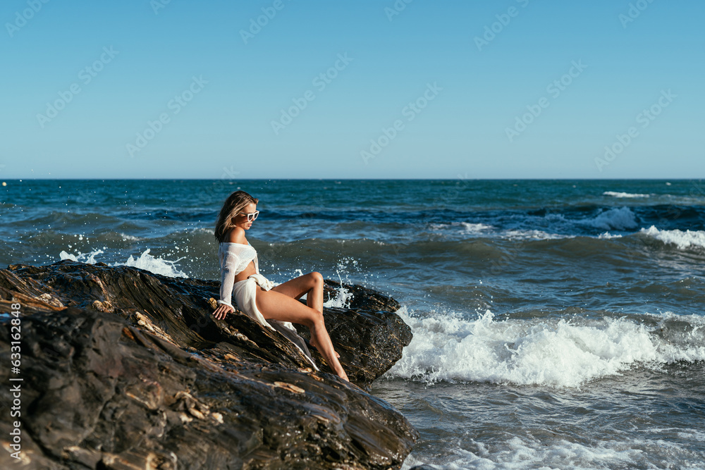 A beautiful girl in white glasses and white clothes sits on the rocks on the seashore