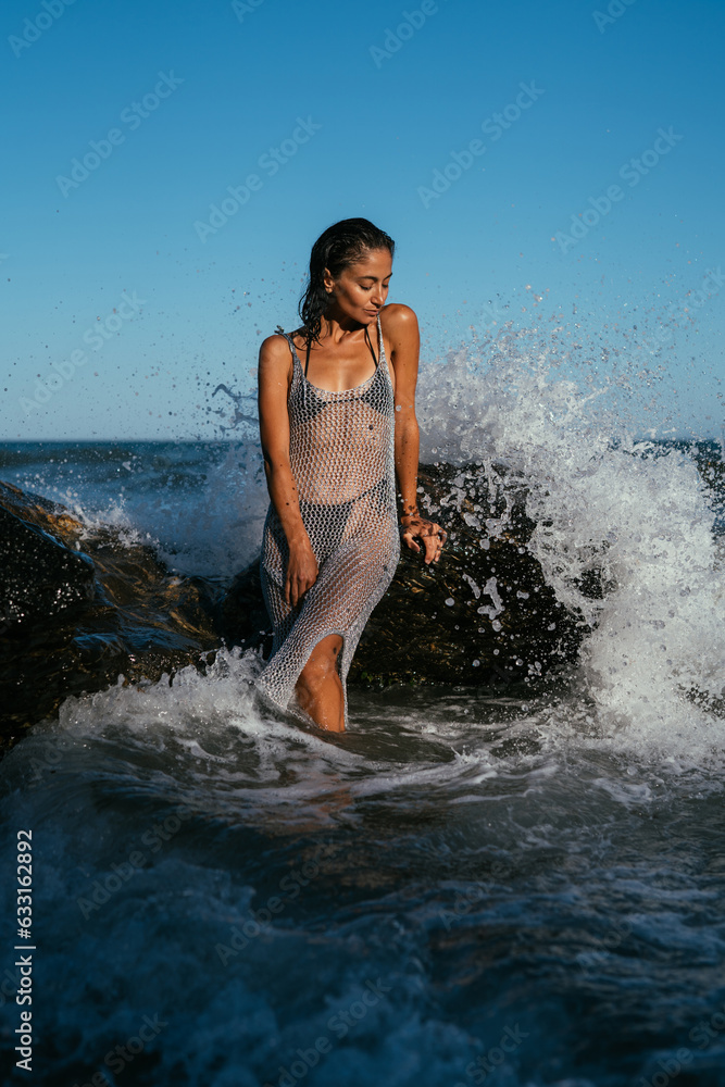 A beautiful wet brunette girl in a silvery net stands in the water on the seashore A beautiful wet brunette girl in a silvery net stands in the water on the seashore