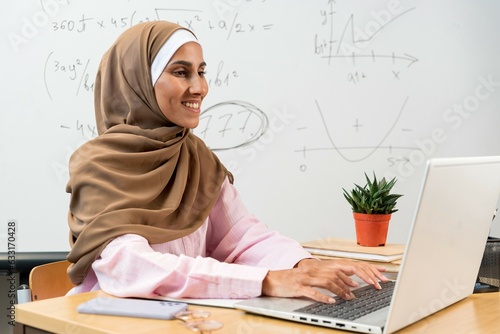 Cheerful Muslim woman studying online on laptop, typing on keyboard, classroom interior. Confident satisfied middle eastern woman in hijab starts new project typing on computer. Education and learning