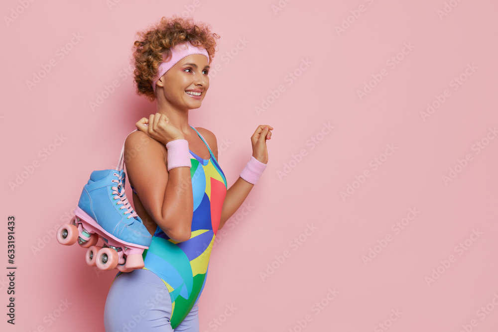 Happy curly girl dressed in colorful leotard stands isolated on pink background with pair of roller skates, sport lifestyle concept, copy space