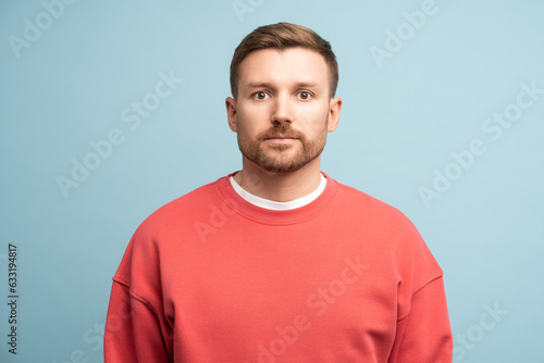 Emotionless middle aged man with serious face expression try calm stands on blue wall studio background. Portrait male sadly looking to camera, thinking about problem, holding back emotions copy space photo