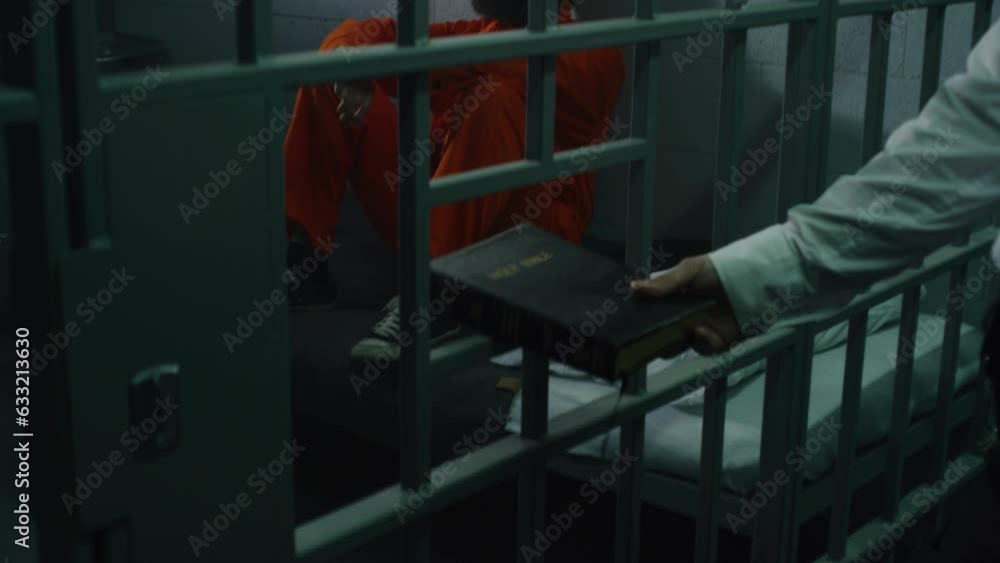 Male prisoner in orange uniform sits on the bed in prison cell. Jailer gives Bible through metal bars. Criminal serves imprisonment term for crime in jail. Detention center or correctional facility. Male prisoner in orange uniform sits on the bed in prison cell. Jailer gives Bible through metal bars. Criminal serves imprisonment term for crime in jail. Detention center or correctional facility.