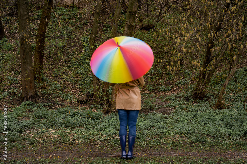 young woman in a beige coat and rain boots stands in forest or park and holds colorful bright rainbow umbrella. Girl spins umbrella for blur effect. Slow shutter speed. Autumn weather in city