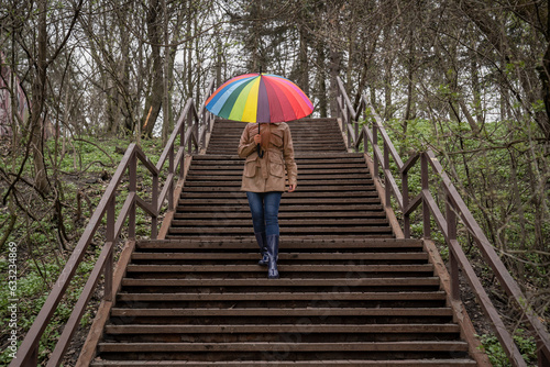 young woman in a beige coat and rain boots stands in forest or park and holds colorful bright rainbow umbrella. Autumn weather in city, girl is walking on a wooden stairs in the park. High stairway