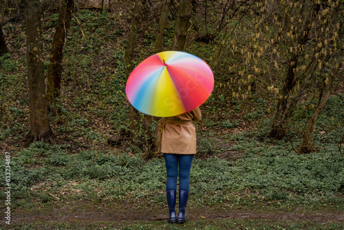 young woman in a beige coat and rain boots stands in forest or park and holds colorful bright rainbow umbrella. Girl spins umbrella for blur effect. Slow shutter speed. Autumn weather in city