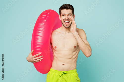 Young man wear green shorts swimsuit relax near hotel pool hold inflatable rubber ring scream sharing hot news about sales isolated on plain blue background. Summer vacation sea rest sun tan concept.
