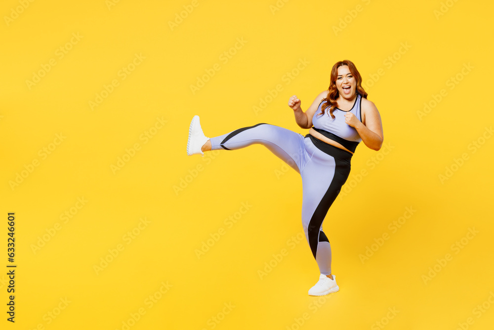 Full body side view young chubby plus size big fat fit woman wears blue top warm up training raise upleg clench fist fight isolated on plain yellow background studio home gym. Workout sport concept.