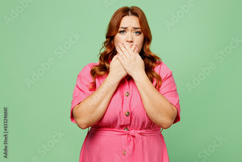 Young scared fearful surprised panicked sad chubby overweight redhead woman wear casual clothes pink dress cover mouth with hand isolated on plain pastel light green color background Lifesyle concept photo