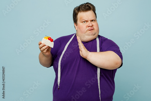 Young confused shocked sad chubby overweight man wear purple t-shirt casual clothes measure tape reject eating cake show stop gesture isolated on plain pastel blue cyan background. Lifestyle concept.