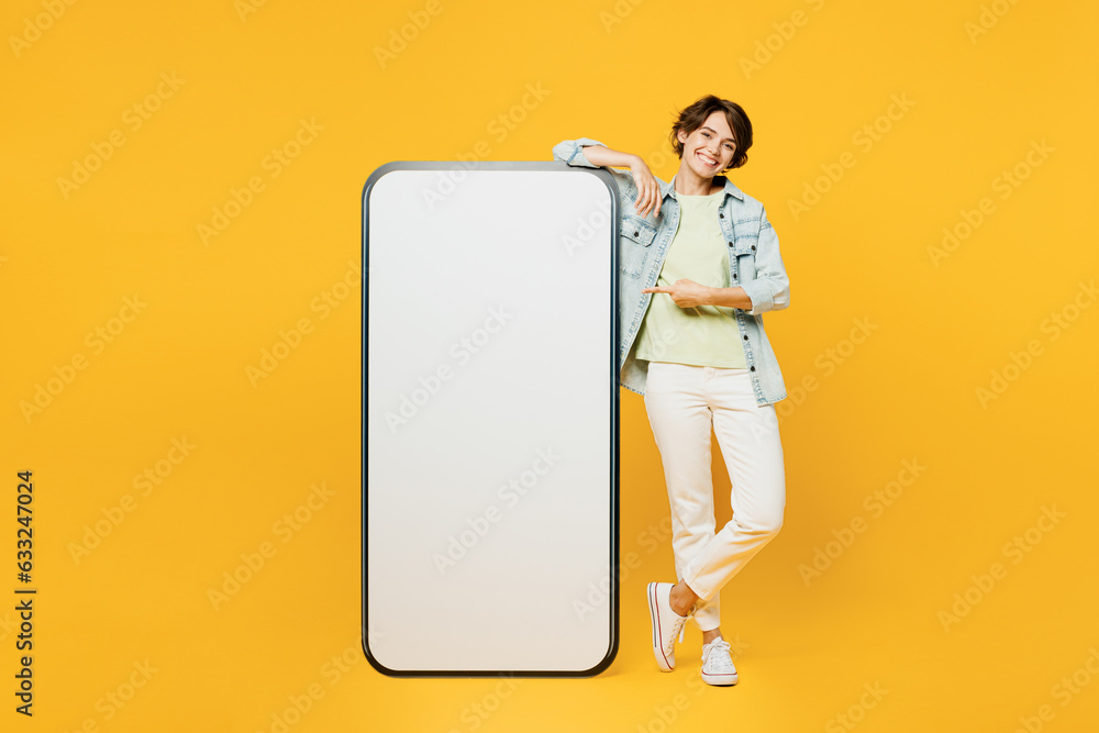 Full body smiling young woman she wear green t-shirt denim shirt casual clothes point index finger on big huge blank screen mobile cell phone smartphone with area isolated on plain yellow background. Full body smiling young woman she wear green t-shirt denim shirt casual clothes point index finger on big huge blank screen mobile cell phone smartphone with area isolated on plain yellow background.