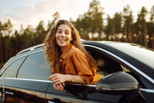 Happy curly woman smiling and enjoying nature leaning out of the car window. Feeling of freedom on the open road. Beauty and travel concept. Active lifestyle.