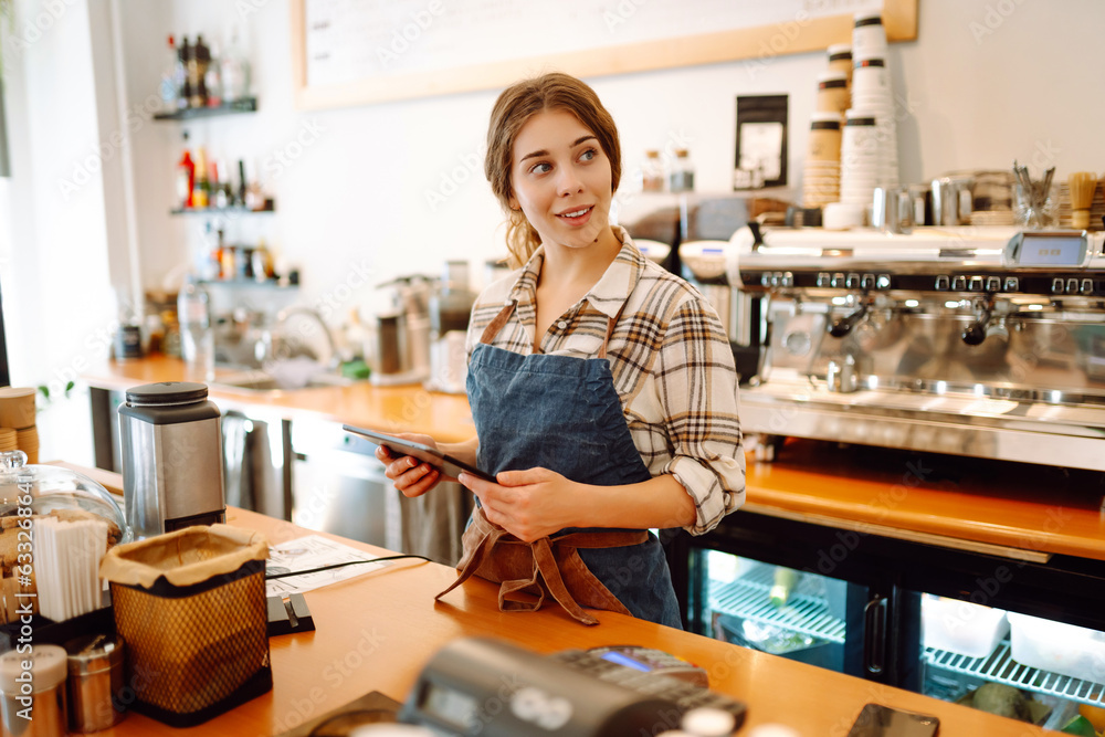Successful small business owner stands behind the counter of coffee shop with digital tablet, takes order. Portrait of beautiful woman barista. Business concept of seller-entrepreneur Successful small business owner stands behind the counter of coffee shop with digital tablet, takes order. Portrait of beautiful woman barista. Business concept of seller-entrepreneur