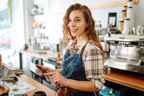 Successful small business owner stands behind the counter of coffee shop with digital tablet, takes order. Portrait of beautiful woman barista. Business concept of seller-entrepreneur