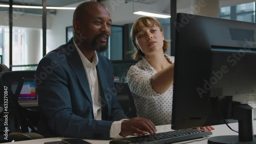 Mature man and young woman in businesswear smiling and talking by computer on desk in office